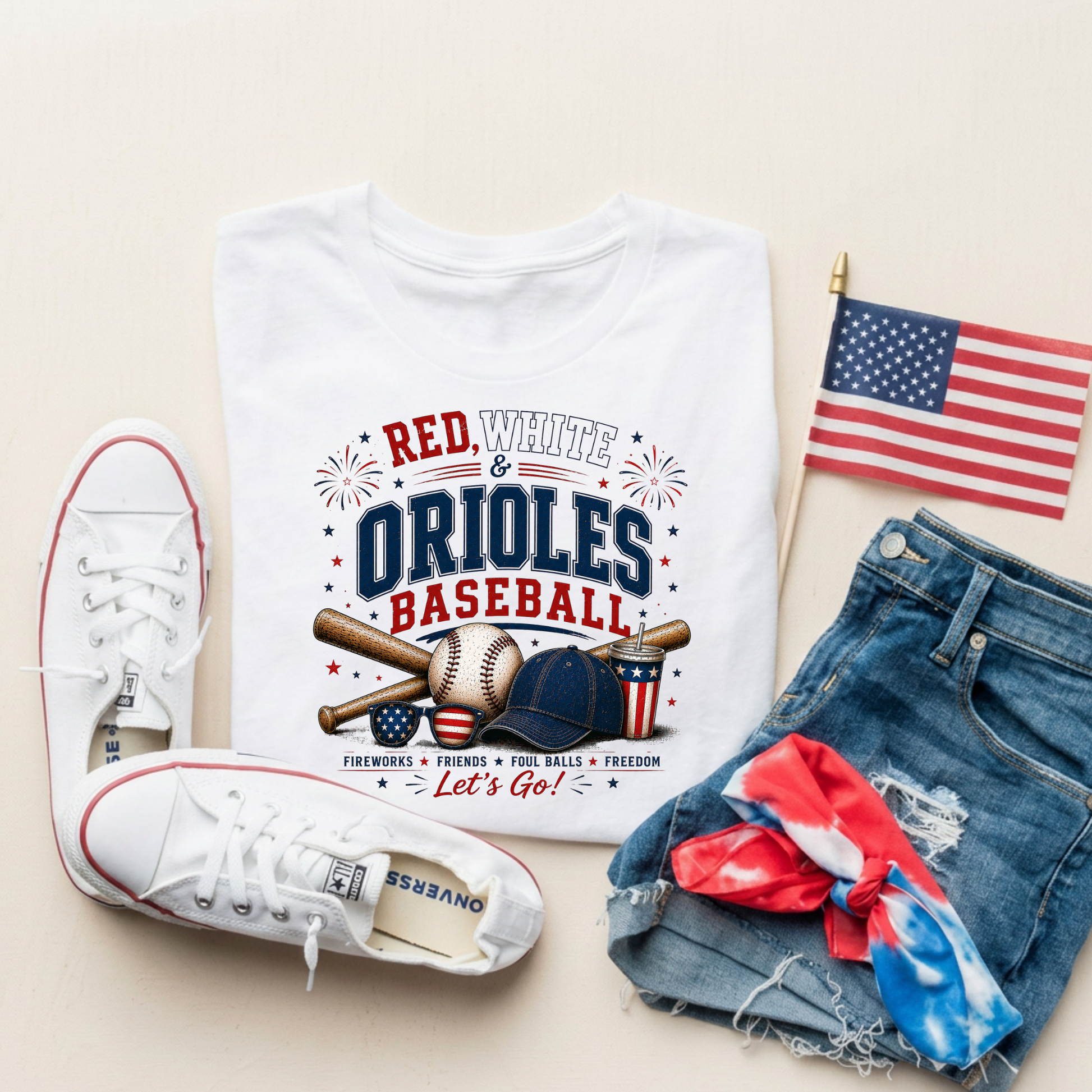 White t-shirt with 'Red, White & Orioles Baseball' design, white sneakers, denim shorts, and American flag on a beige background.