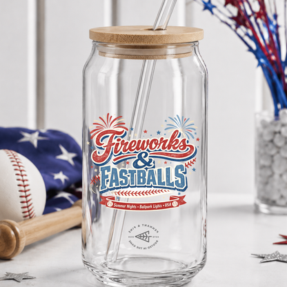 Clear glass tumbler with wooden lid and straw, featuring 'Fireworks & Fastballs' design, on a table with American flag and baseball.